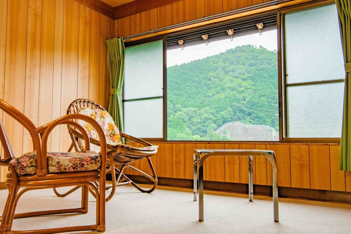 A room with wooden walls, two wicker chairs with floral upholstery, a small table, and a large window with a view of a forested hill.