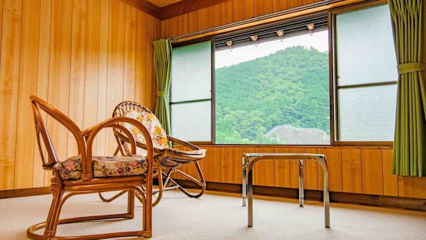 A room with wooden walls, two wicker chairs with floral upholstery, a small table, and a large window with a view of a forested hill. - Minshuku Yaichi (Gujo)
