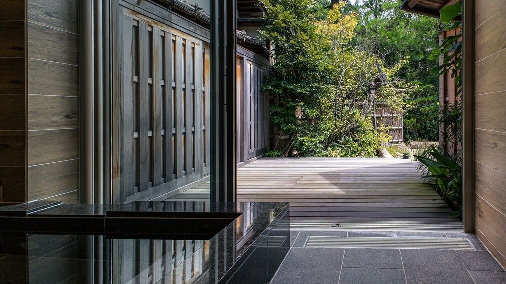 Ein traditionelles japanisches Gebäude mit Holzbeschichtung, einem mit Fliesen belegten Weg, der zu einer Holztür führt, und einem Garten mit Bäumen und Sträuchern.