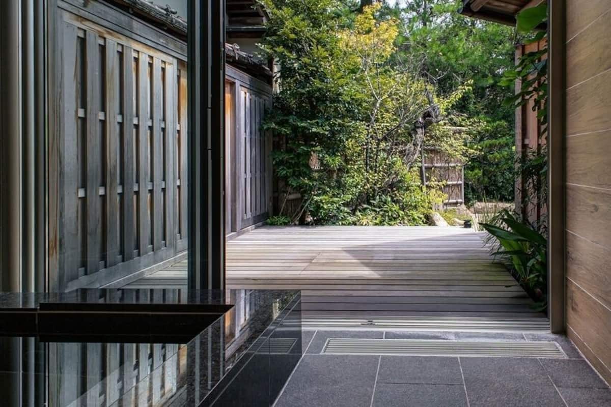 A traditional Japanese-style building with a wooden exterior, a tiled pathway leading to a wooden door, and a garden with trees and shrubs.