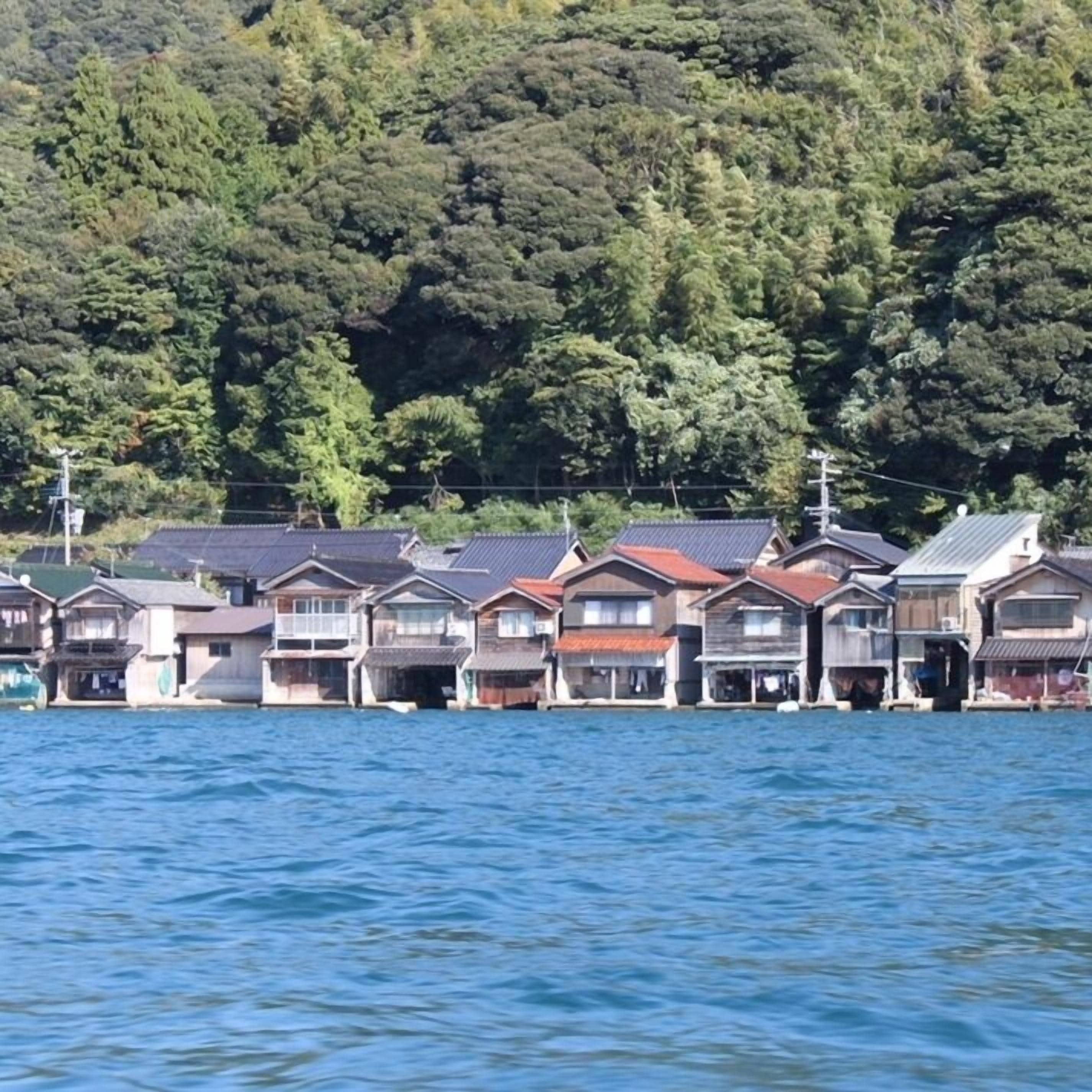 Uma fileira de casas japonesas tradicionais à beira-mar, com uma encosta arborizada ao fundo.