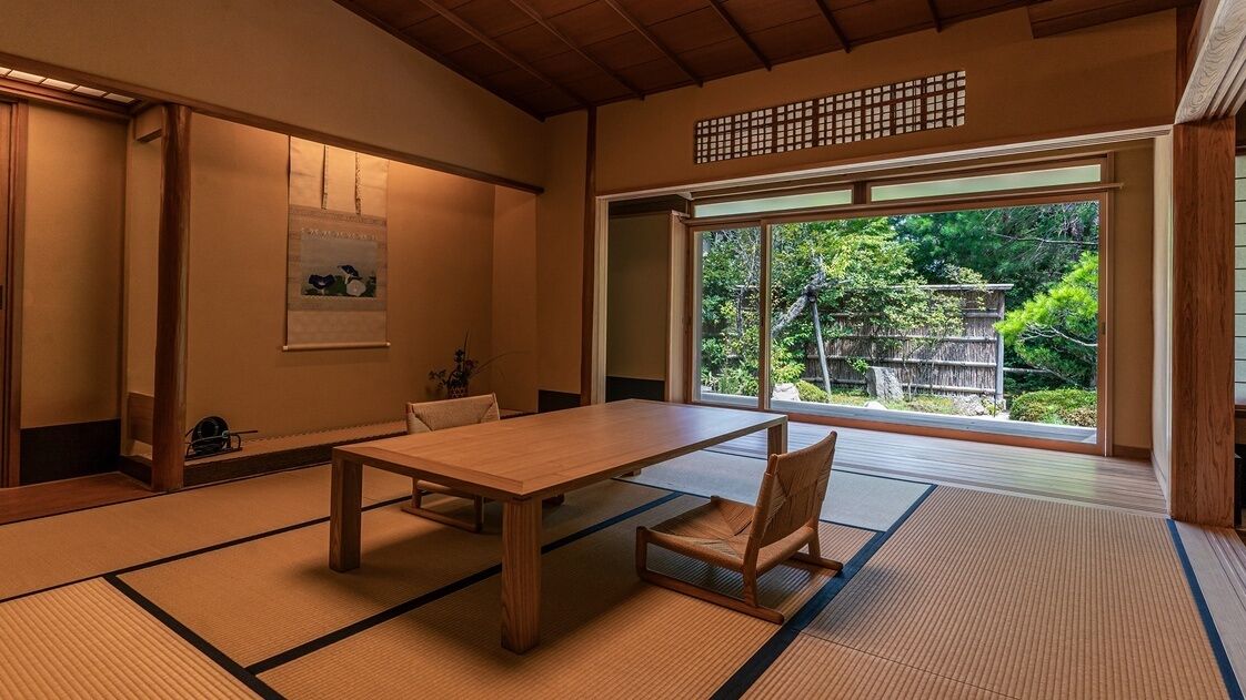 Ein traditioneller japanischer Raum mit Tatami-Boden, einem Holztisch, einem Stuhl und einem großen Fenster mit Blick auf Grünflächen.