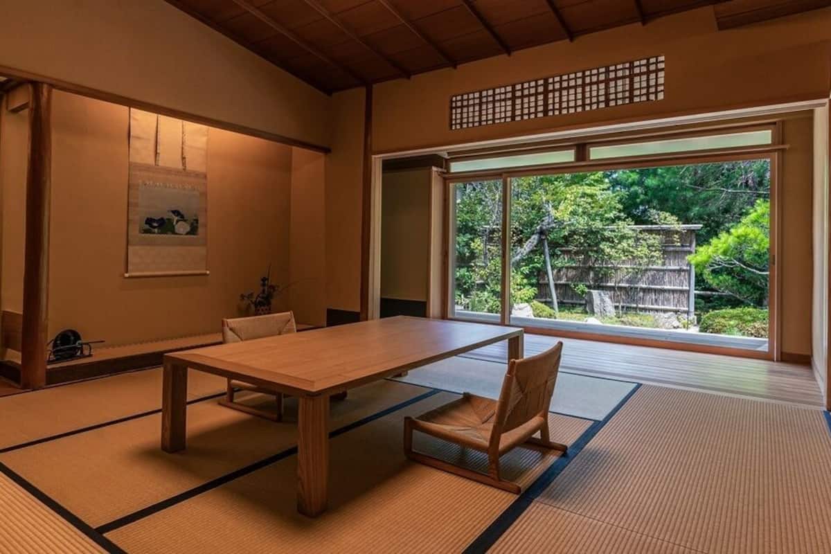A traditional Japanese room with tatami flooring, a wooden table, a chair, and a large window with a view of greenery.