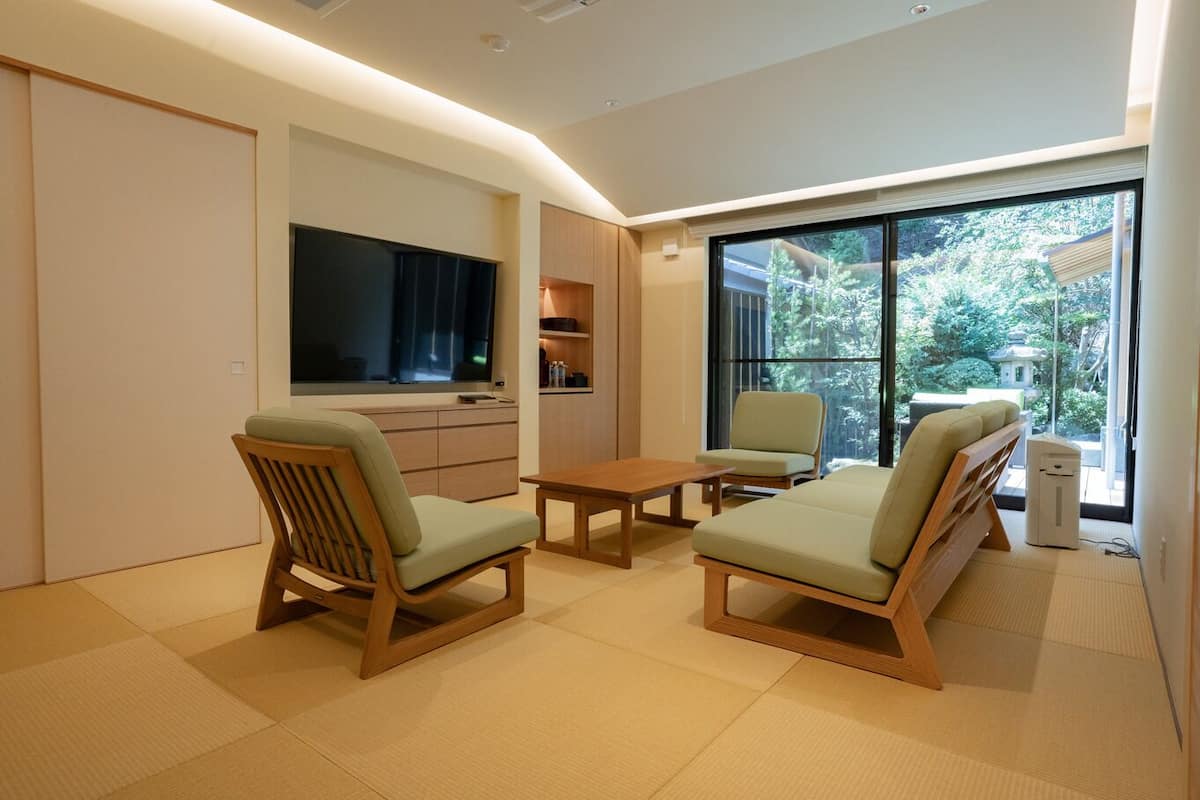 A modern living room with tatami flooring, a flat-screen TV, wooden furniture, and a view of greenery outside.
