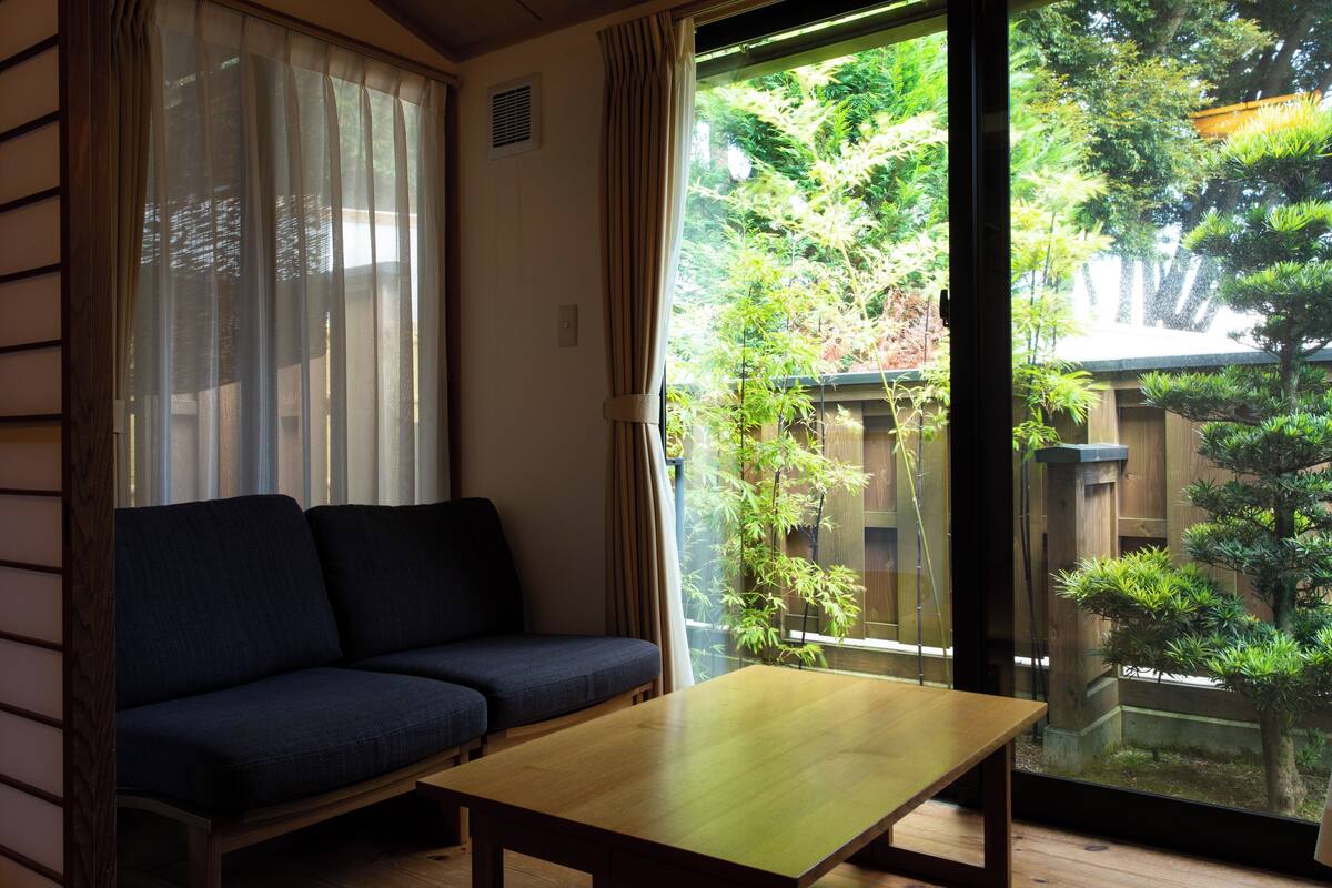 A room with a sofa, a table, and a view of a garden through a glass door.