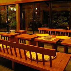 An outdoor seating area with wooden tables and benches, potted plants, and a view of a building with large windows. - Kamai Onsen Ippenan (Kyotango)