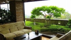 A room with a sofa, a coffee table, and a view of a garden with a tree and stone wall. - Dake Onsen Nagame no Yakata Kounkaku (Nihommatsu)