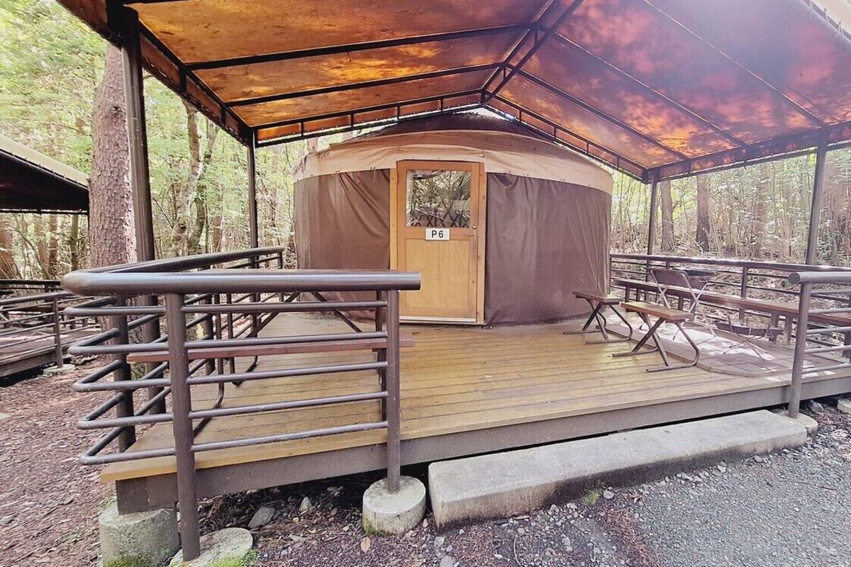 Une structure circulaire en bois, dotée d’une porte portant l’inscription « P6 » et d’une terrasse en bois couverte, aménagée avec des bancs.