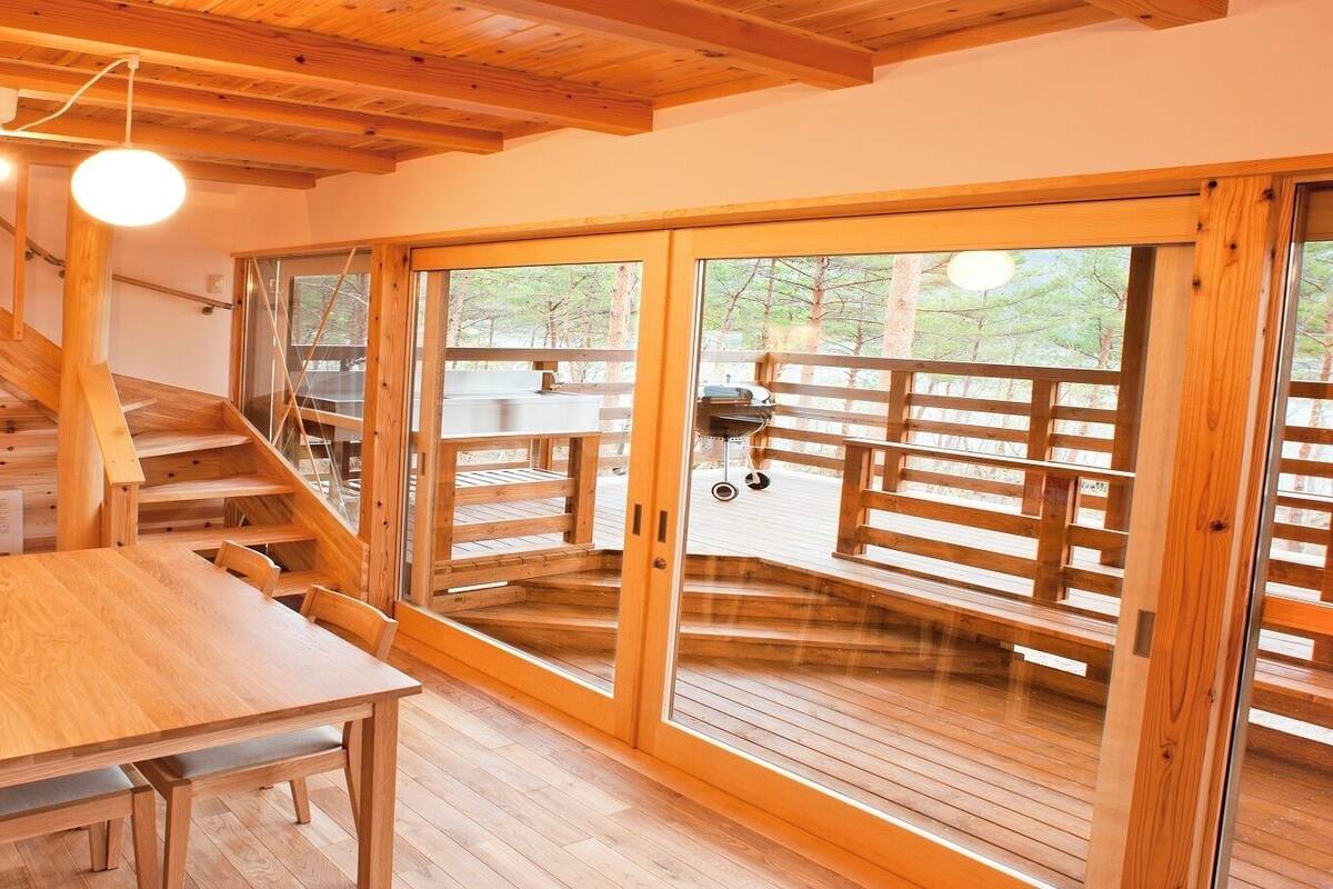Un intérieur en bois avec un escalier, une table et de grandes portes-fenêtres coulissantes donnant sur une terrasse extérieure.