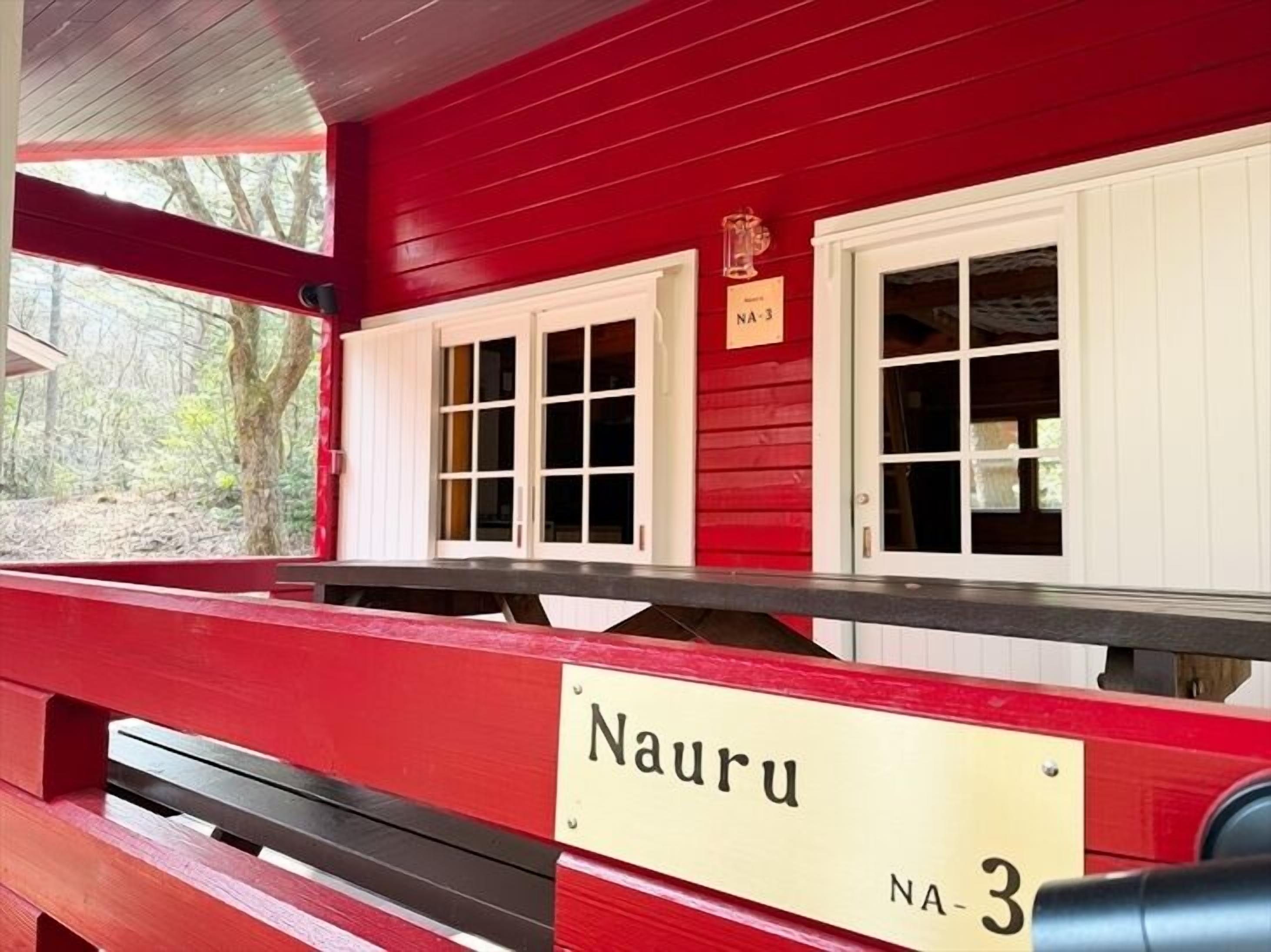 Un edificio rosso con un cartello che riporta 'Nauru NA-3' e due finestre con persiane bianche.