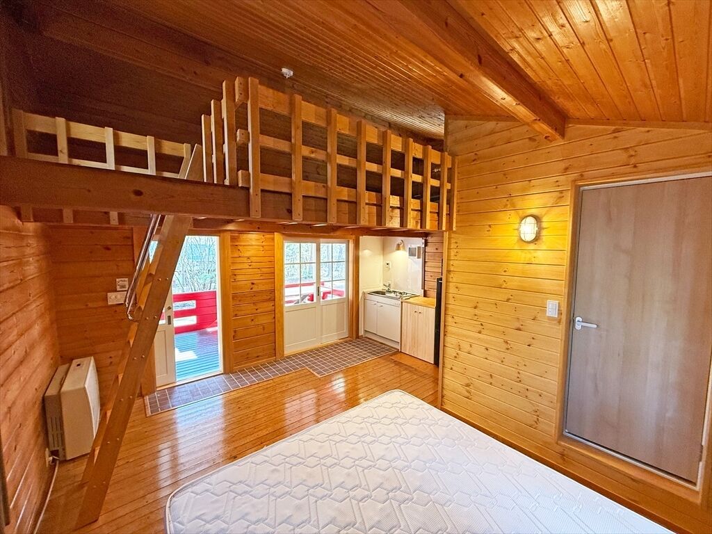 A wooden cabin interior with a loft bedroom, a kitchen area, and a large door.