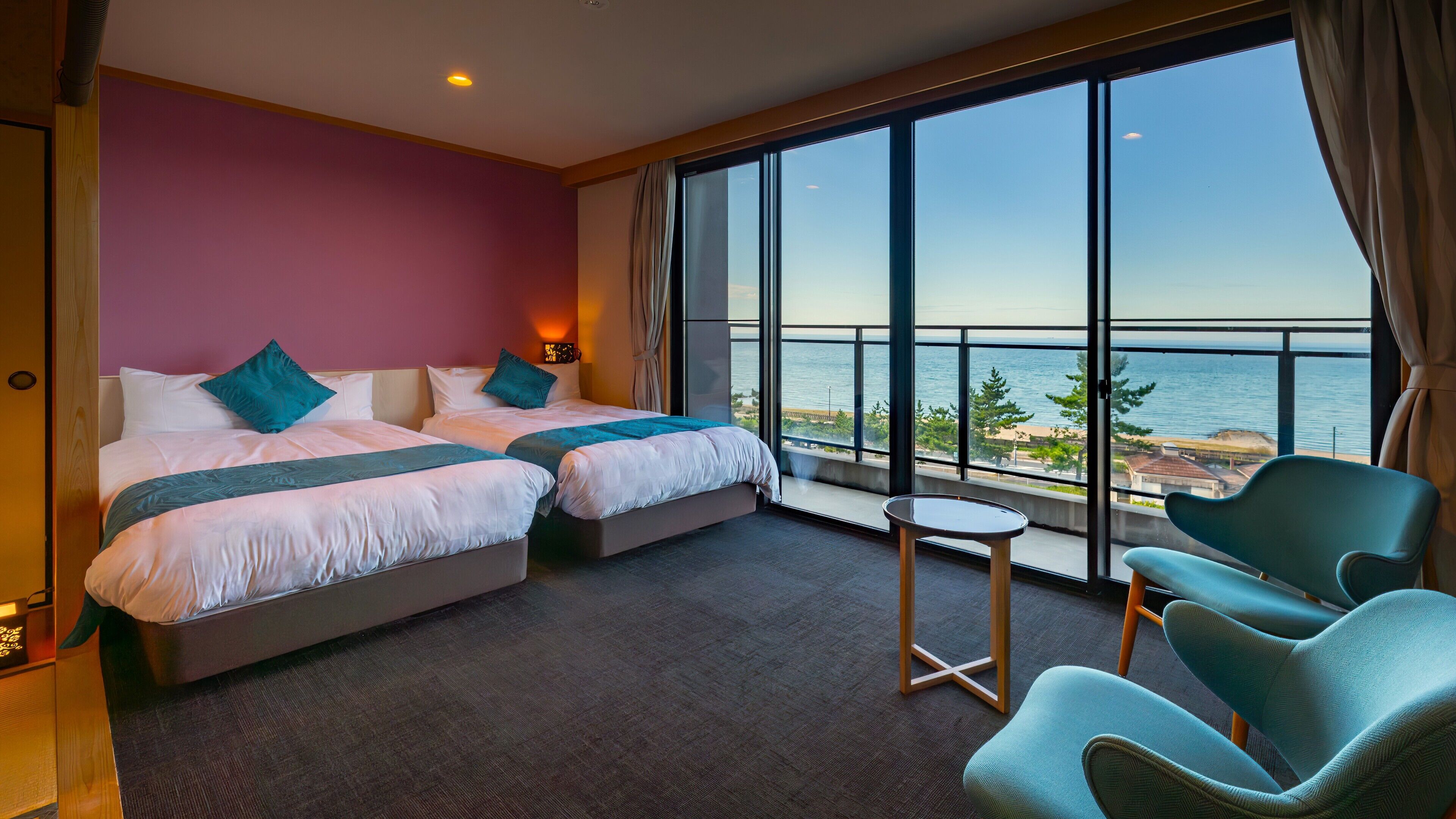 A hotel room with two beds, a small table, two chairs, a large window with a view of the sea, and a ceiling light.