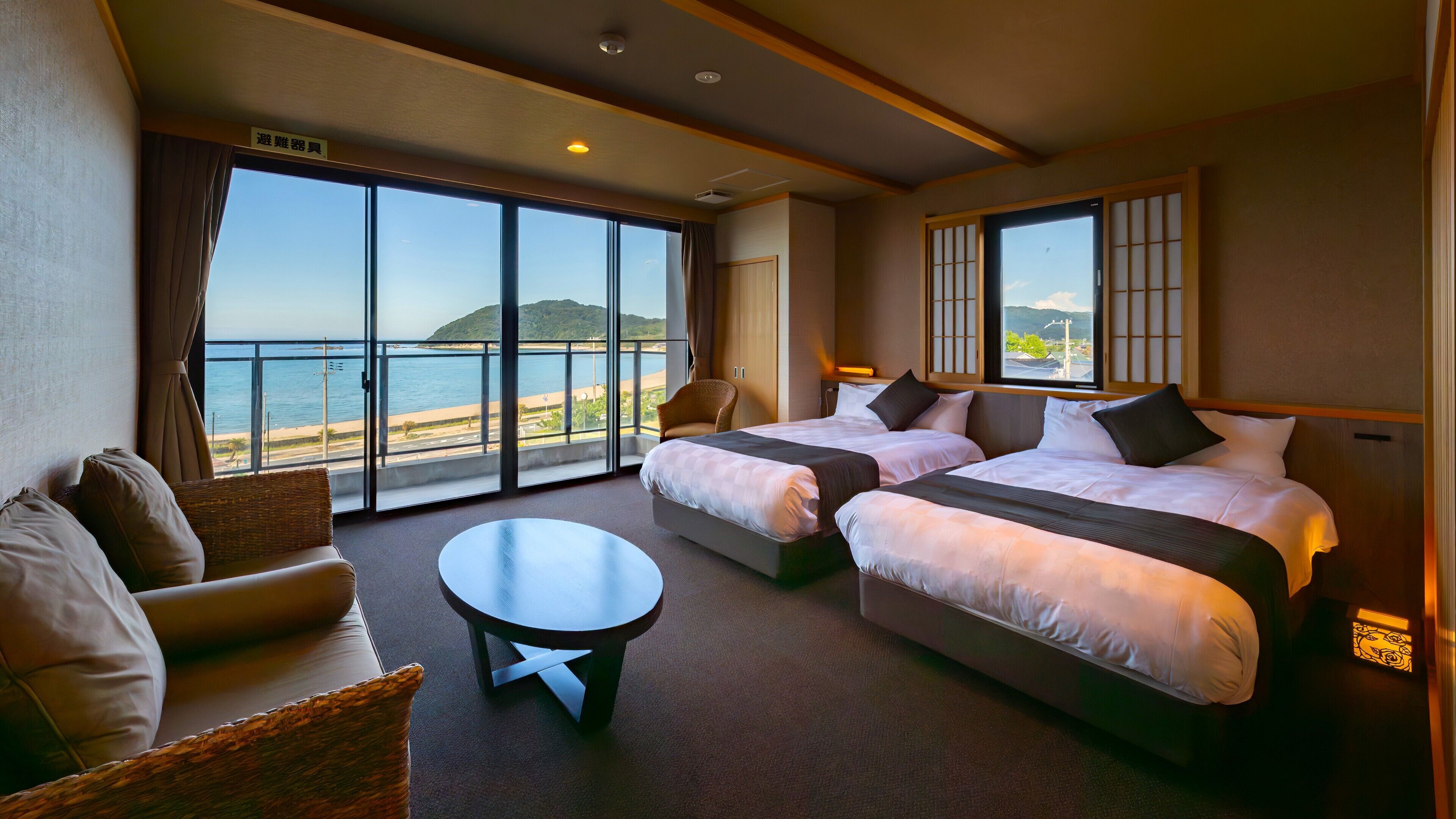 A hotel room with two beds, a small table, a chair, and large windows offering a view of the sea and a distant hill.