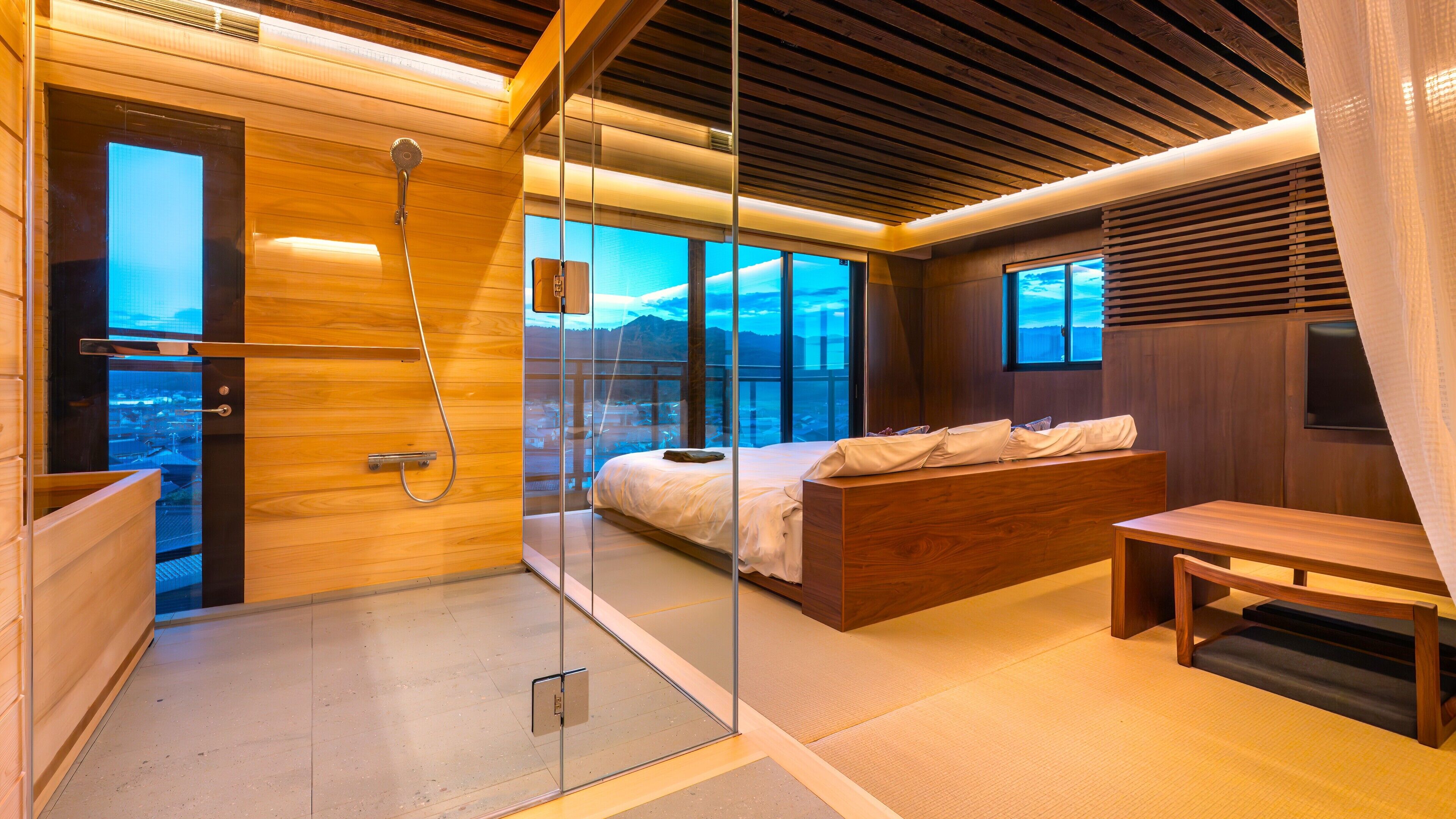 A modern bedroom with a glass shower enclosure, a bed with a wooden headboard, and a view of the cityscape.