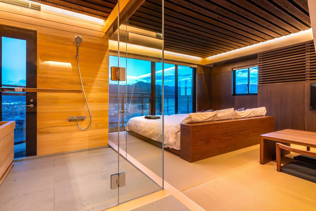 Camera da letto moderna con box doccia in vetro, letto con testiera in legno e vista sulla città.