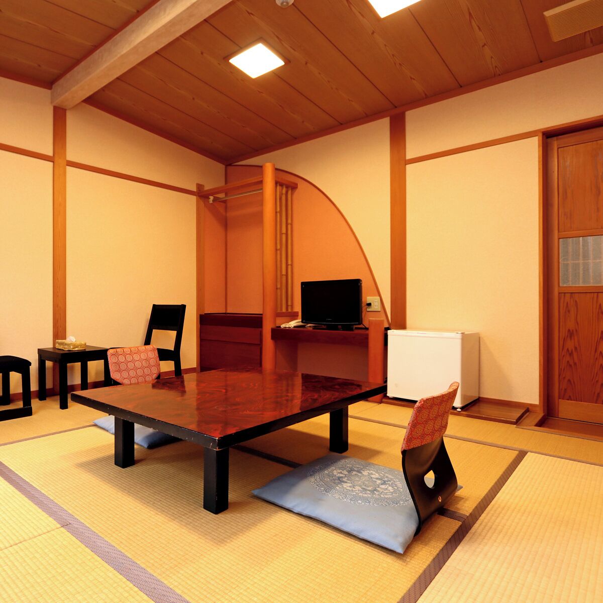 Une pièce avec un sol recouvert de tatamis, une petite table, des chaises, un téléviseur et un radiateur.
