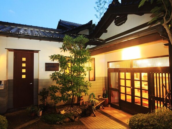 A traditional Japanese building with a tiled entrance, a wooden door, and a well-maintained garden with various plants.