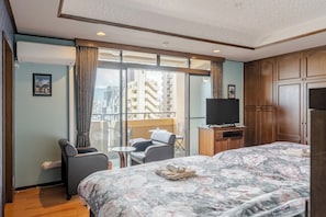 Habitación de hotel con cama, televisor, balcón con una silla y vista a edificios.