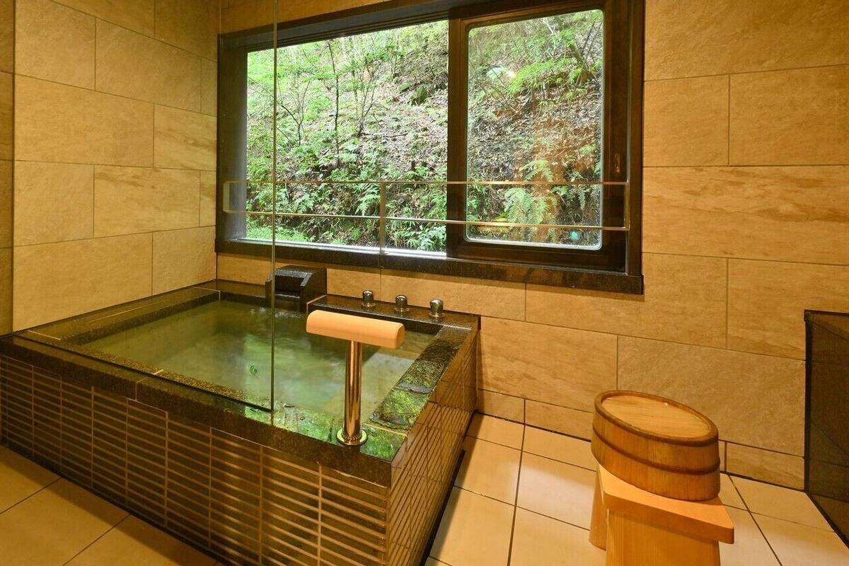 Un cuarto de spa con un jacuzzi envuelto en vidrio, un taburete de madera y un ventanal con vistas al bosque.