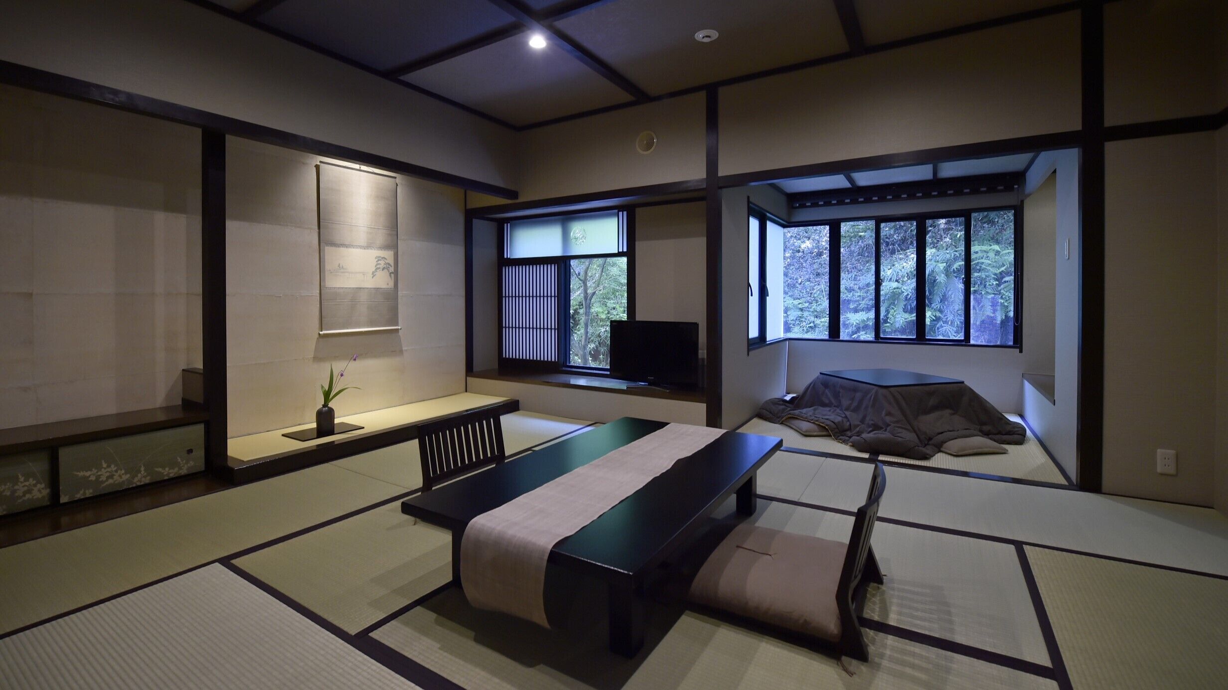 Perinteinen japanilaistyylinen huone, jossa on tatamimatto, matala pöytä, futon-patjalla varustettu sänky, televisio ja ikkuna, josta avautuu näkymä puille.