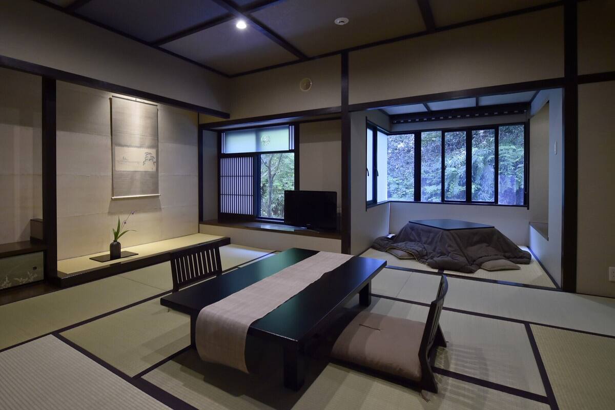 Una habitación tradicional japonesa con suelo de tatami, una mesa baja, una cama con futón, un televisor y una ventana con vistas a árboles.