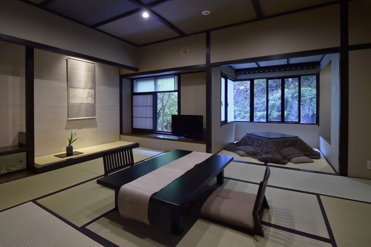 Perinteinen japanilaistyylinen huone, jossa on tatamimatto, matala pöytä, futon-patjalla varustettu sänky, televisio ja ikkuna, josta avautuu näkymä puille.