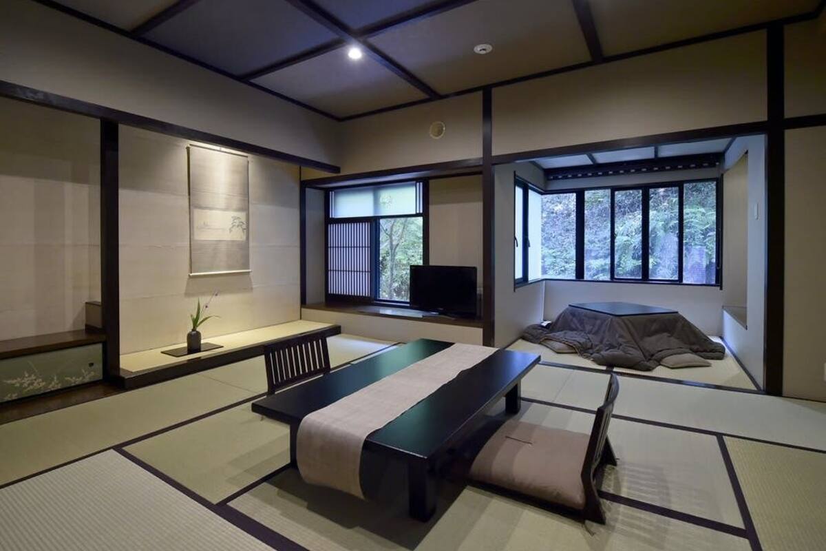 Una habitación de estilo japonés tradicional con suelo de tatami, futón de tatami, una mesa baja, televisión y vistas al exterior a través de amplios ventanales.