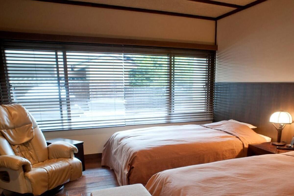 Habitación de hotel con dos camas, una silla de masaje, una mesita de noche con lámpara y una ventana con persianas.