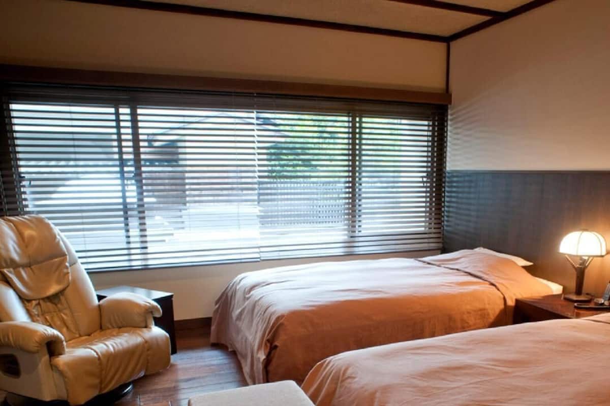 A hotel room with two beds, a massage chair, a bedside table with a lamp, and a window with blinds.