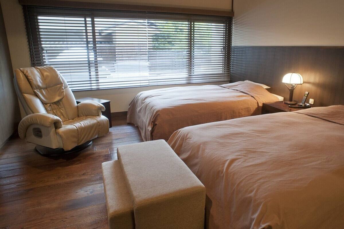Habitación de hotel con dos camas, una silla de masajes, una mesita de noche con lámpara y una ventana con persianas.