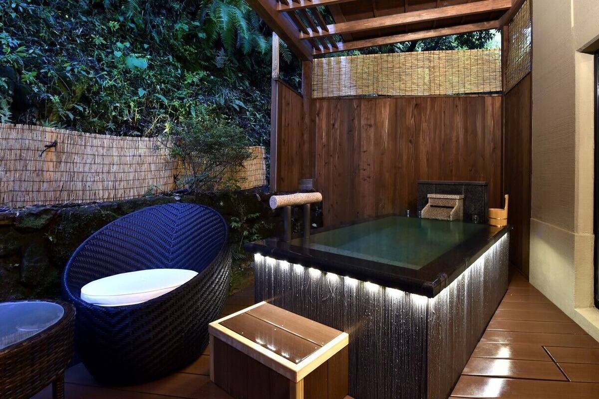 Un jacuzzi con pared de madera y cerramiento de vidrio, rodeado de mobiliario de mimbre y suelo de baldosas.