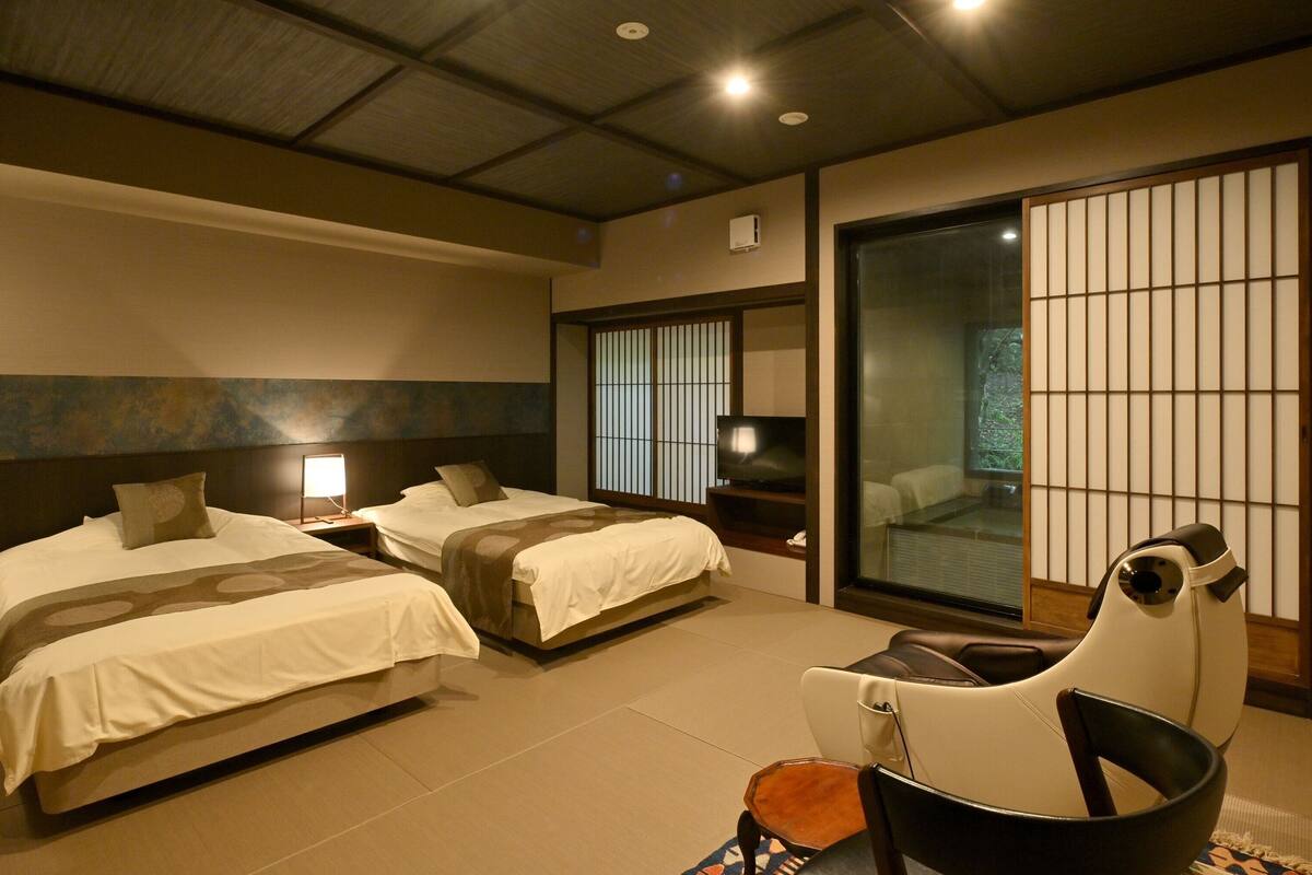 Habitación de hotel con dos camas, televisión, puerta corrediza de vidrio y sillón de masaje.