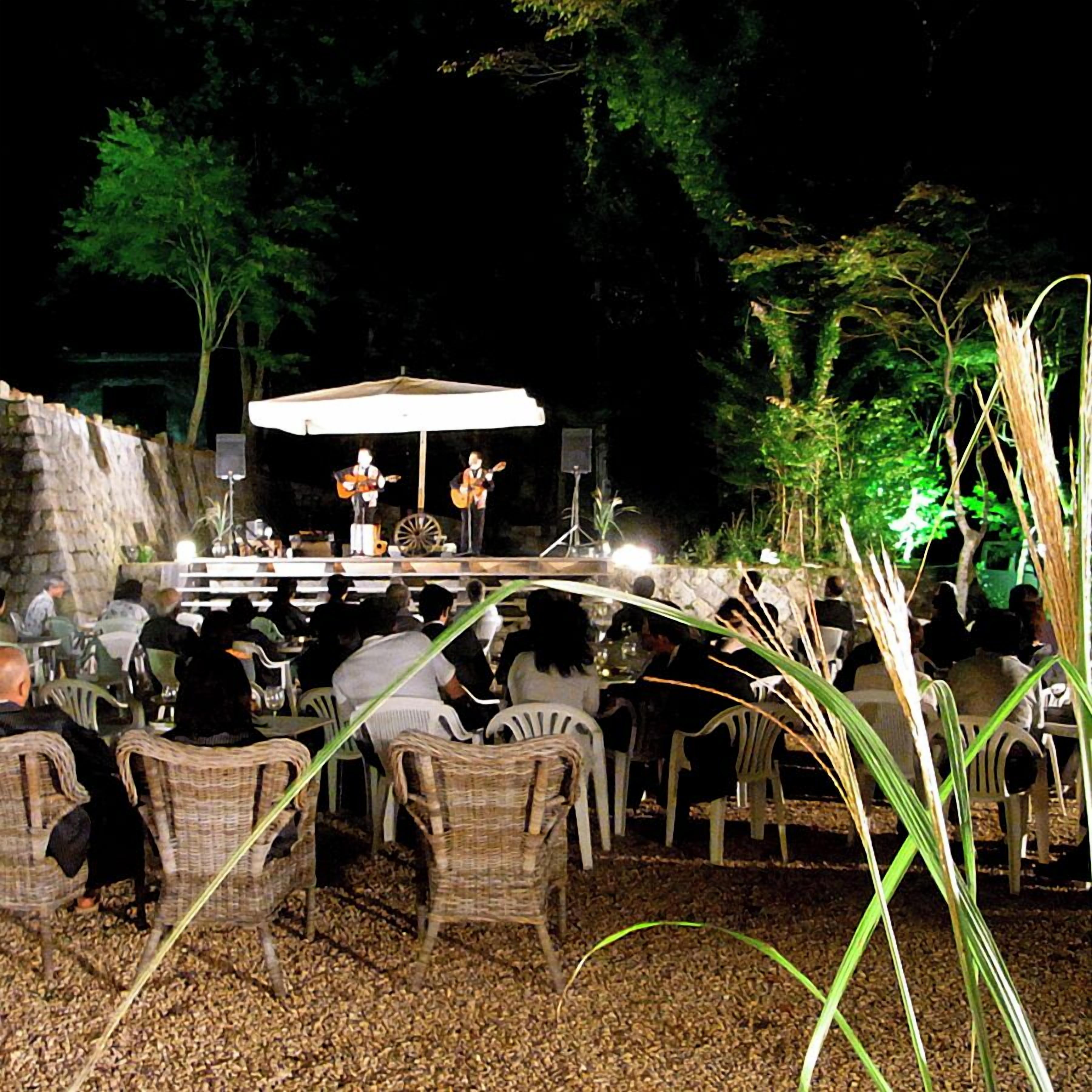 Un espace extérieur aménagé pour les repas, avec des chaises en osier, une scène où se produisent des musiciens et un mur en pierre.