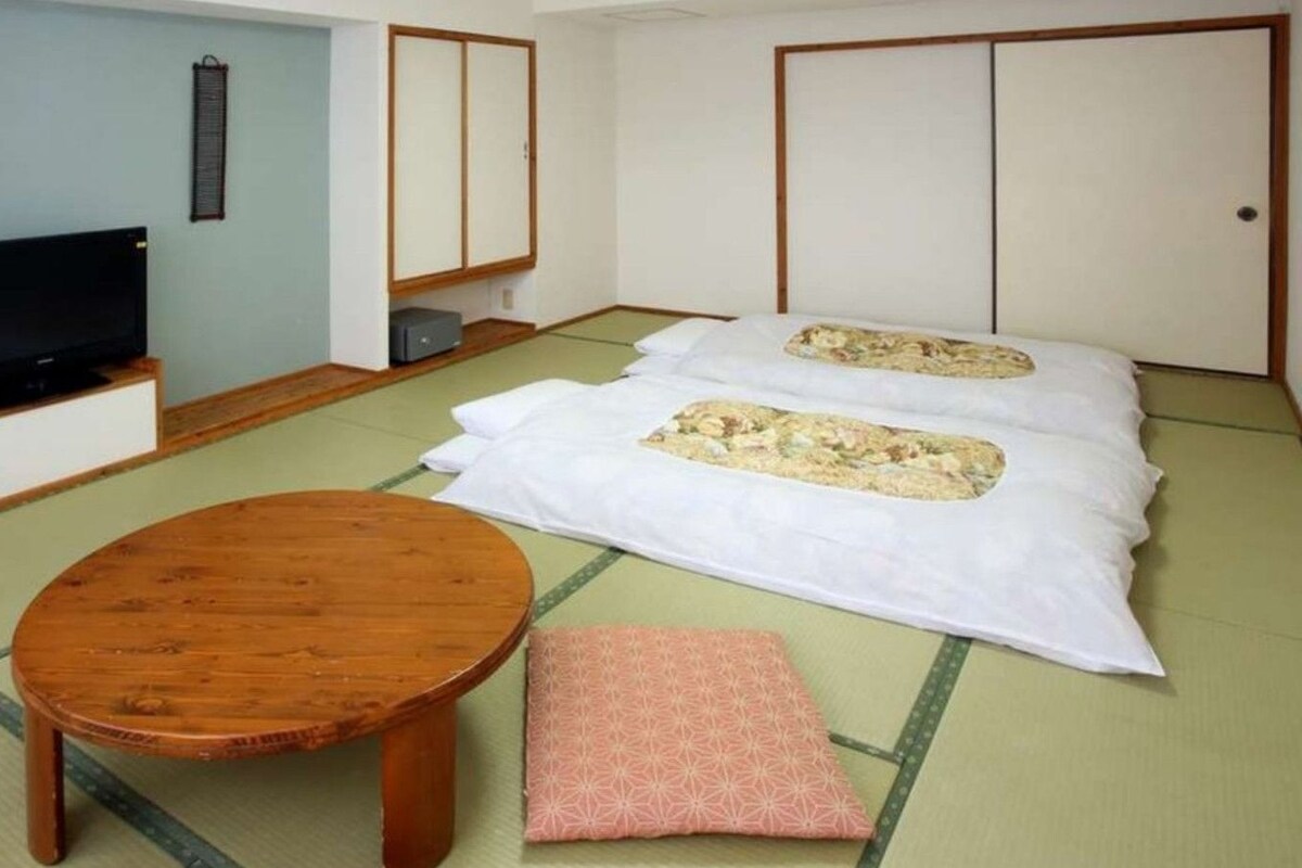 Una habitación japonesa tradicional con suelo de tatami, una mesa de madera baja, un televisor y un futón con almohadas.