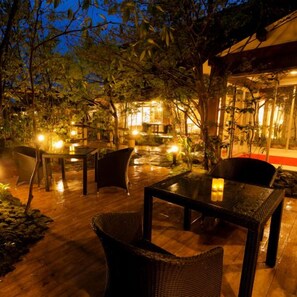 An outdoor patio area with tables and chairs, surrounded by trees and a building with illuminated windows. - Asouchinomaki Onsen Yumeoiso (Aso)