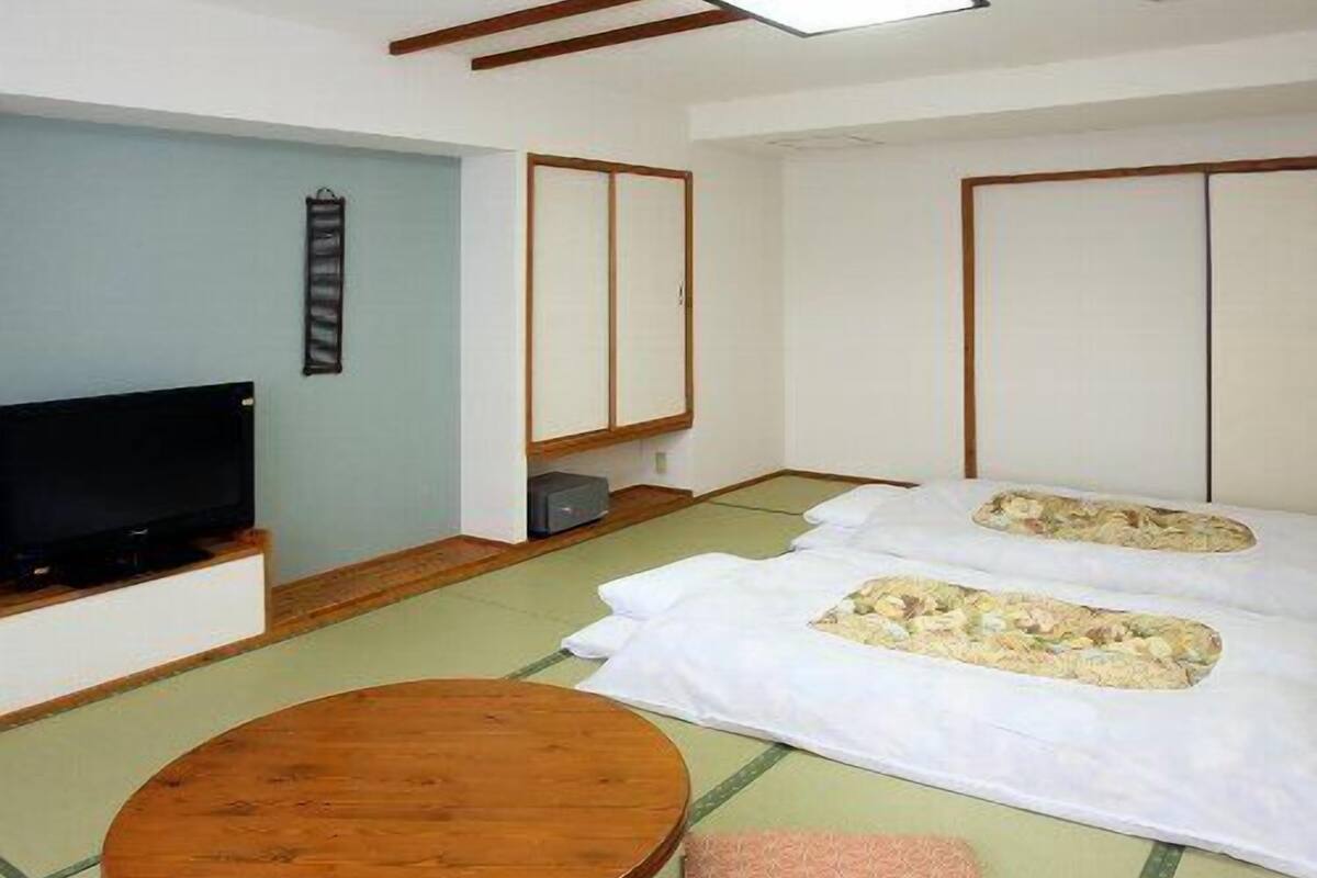 A traditional Japanese-style room with tatami flooring, a television, a wooden table, and futon beds.
