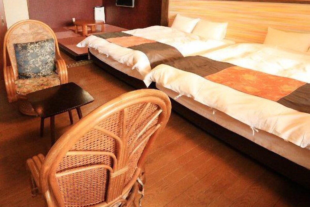 Una habitación con cama, dos sillas, una mesita y televisión montada en la pared.