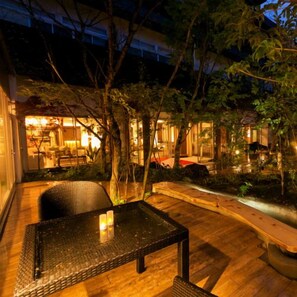 An outdoor patio area with a table and chairs, surrounded by trees and a building with illuminated windows. - Asouchinomaki Onsen Yumeoiso (Aso)