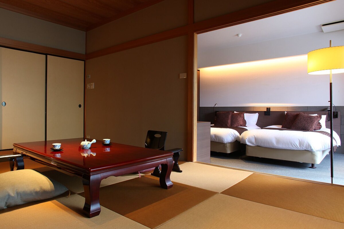 Une pièce de style japonais traditionnel, avec un sol recouvert de tatamis, une basse table en bois, un canapé et un lit doté d’une tête de lit.
