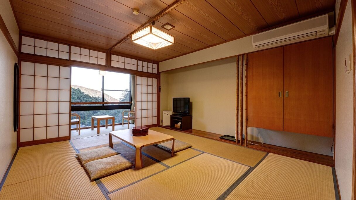 Ein traditioneller japanischer Raum mit Tatami-Boden, einem niedrigen Tisch, einem Fernseher und einer Schiebetür, die auf einen Balkon führt.