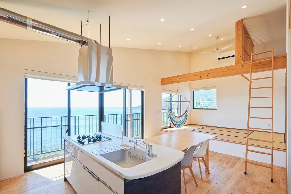 A modern kitchen with a large island, a gas stove, and a sink. There is a balcony with a view of the sea.