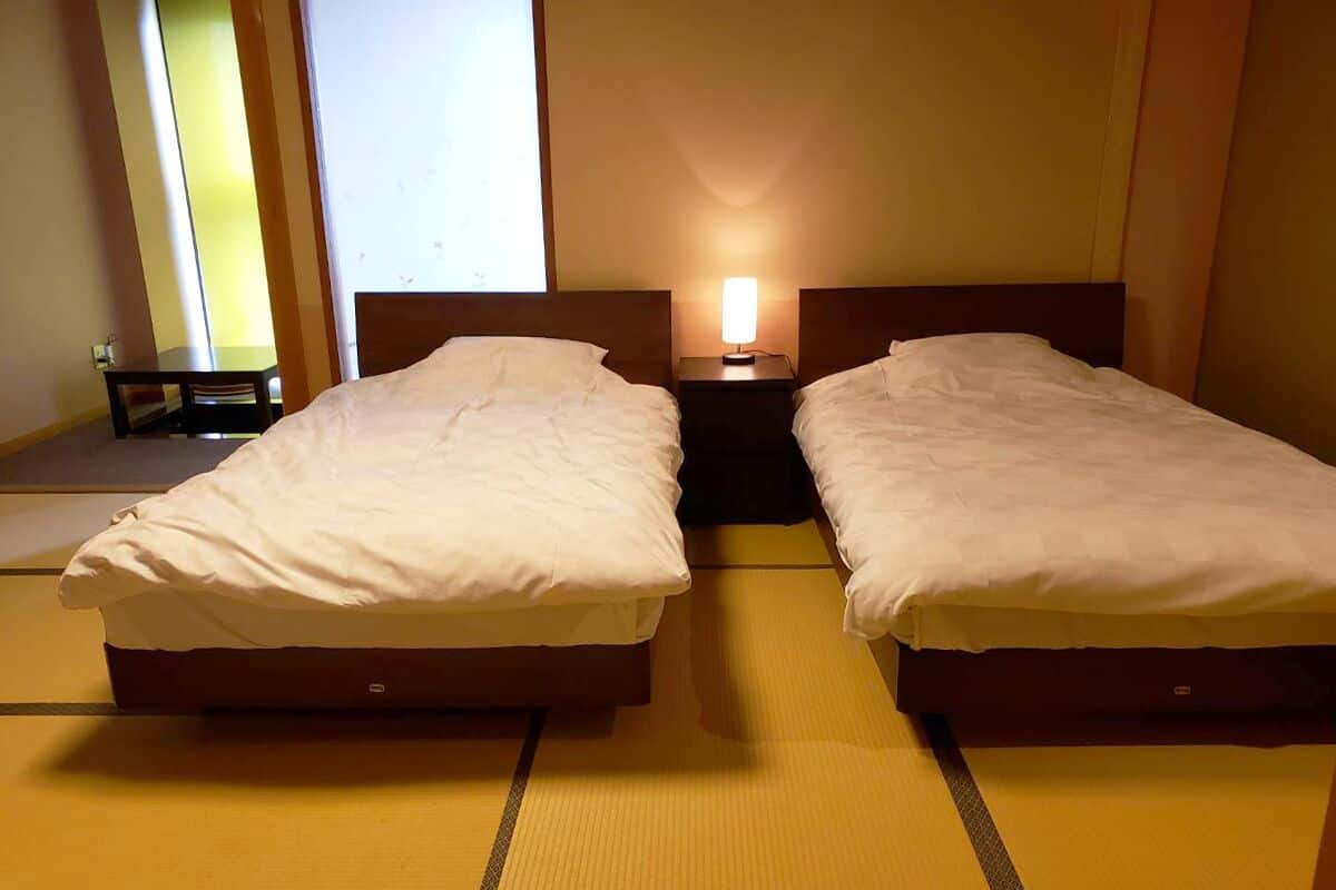A room with two beds, a bedside table with a lamp, and a window.