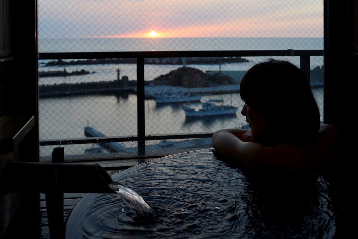 A person relaxing by a window overlooking a sunset view with boats on the water.