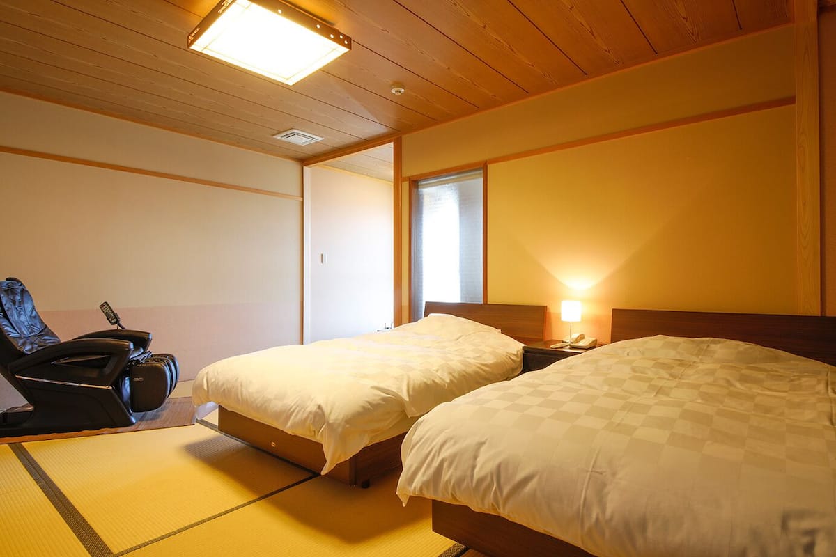 A room with two beds, a massage chair, a bedside table with a lamp, and a window.
