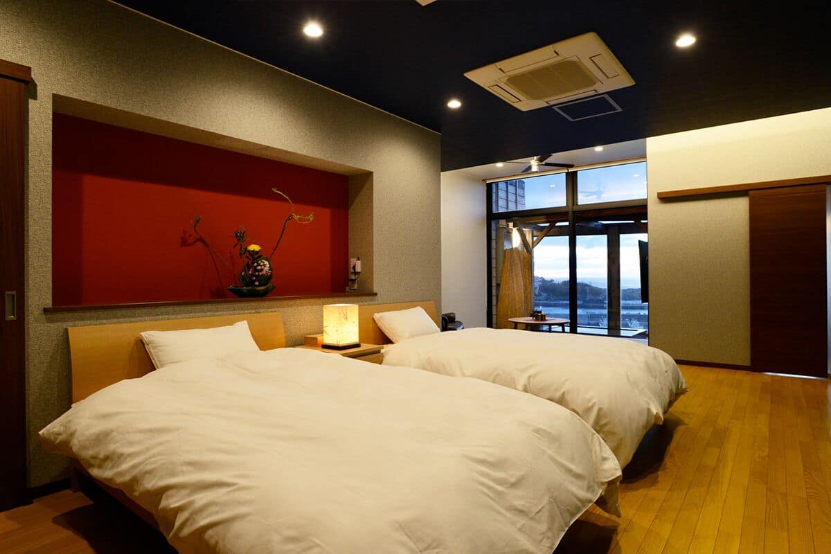 A hotel room with two beds, a red accent wall, a large window, and a view of the outside.