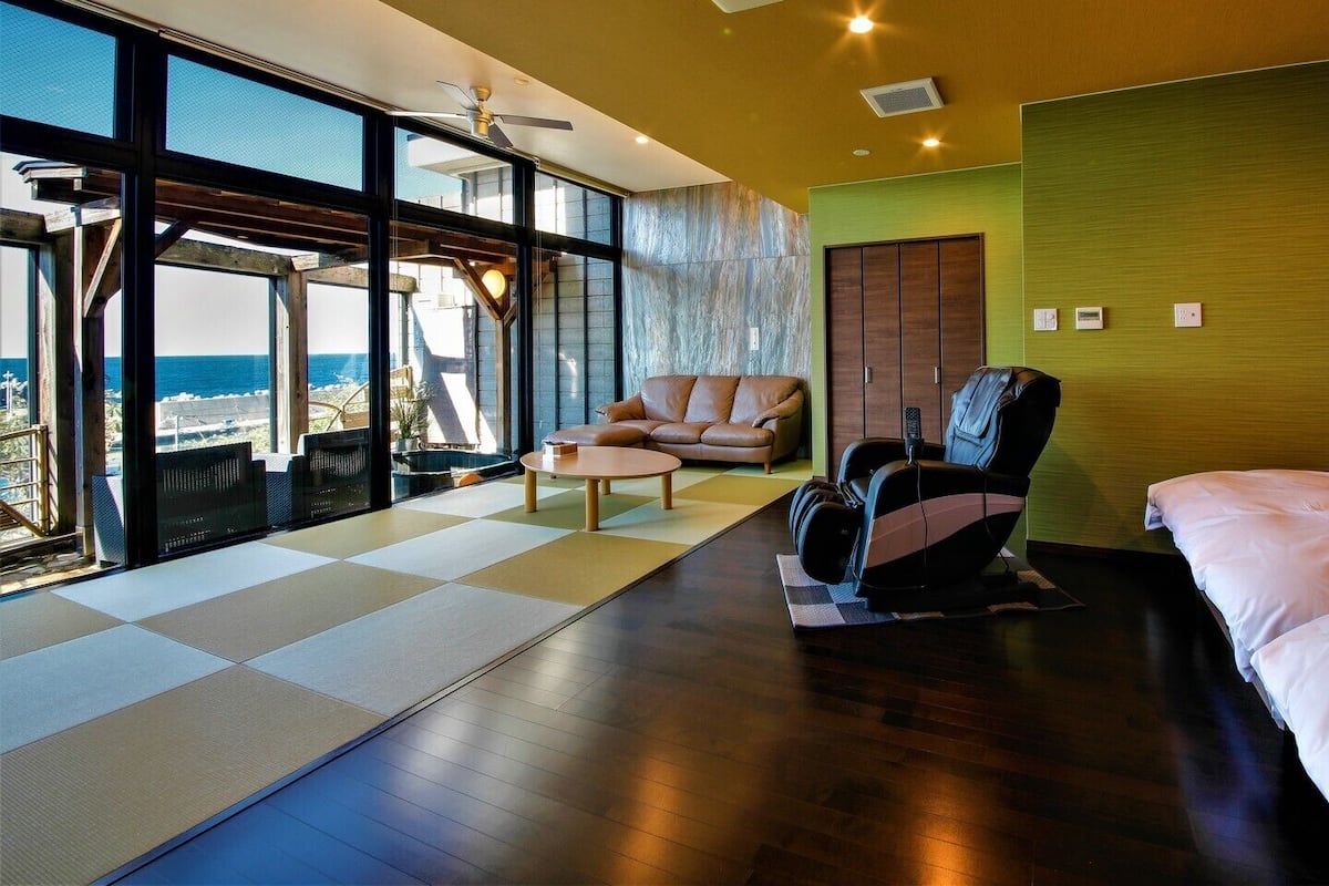 A modern hotel room with a large window, a massage chair, a sofa, and a bed.