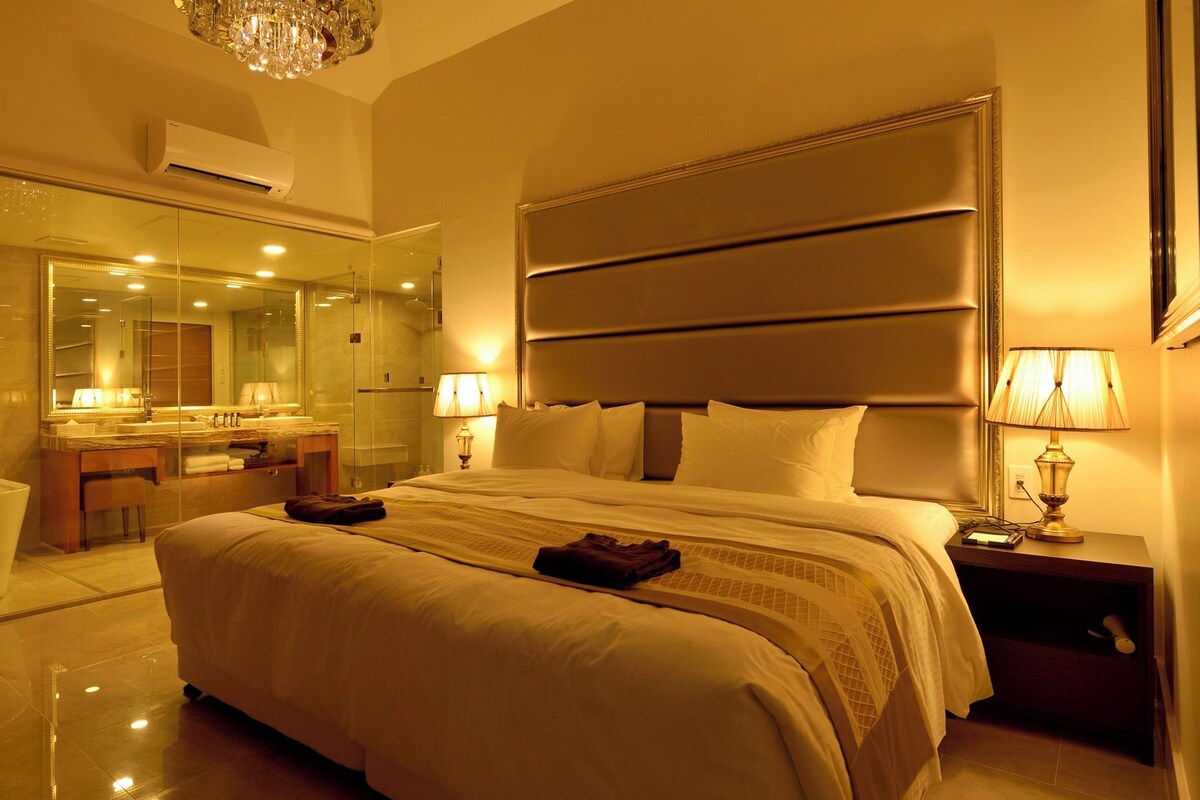 A hotel room with a large bed, a bathroom with a glass shower enclosure, a vanity with a mirror, and a chandelier.