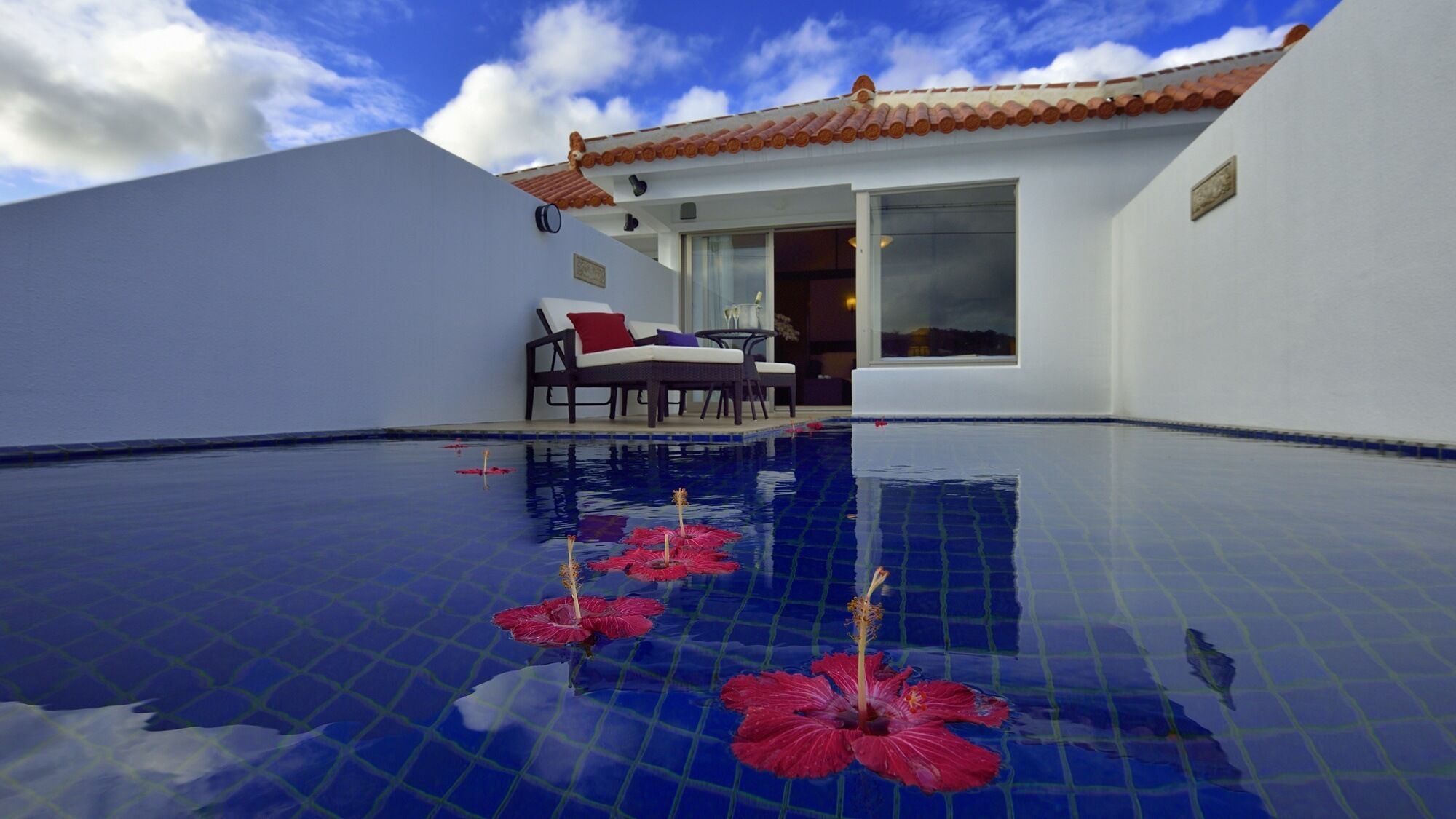 Une piscine bordée de fleurs d’hibiscus, un bâtiment blanc avec un toit en tuiles et une chaise placée à l’extérieur.