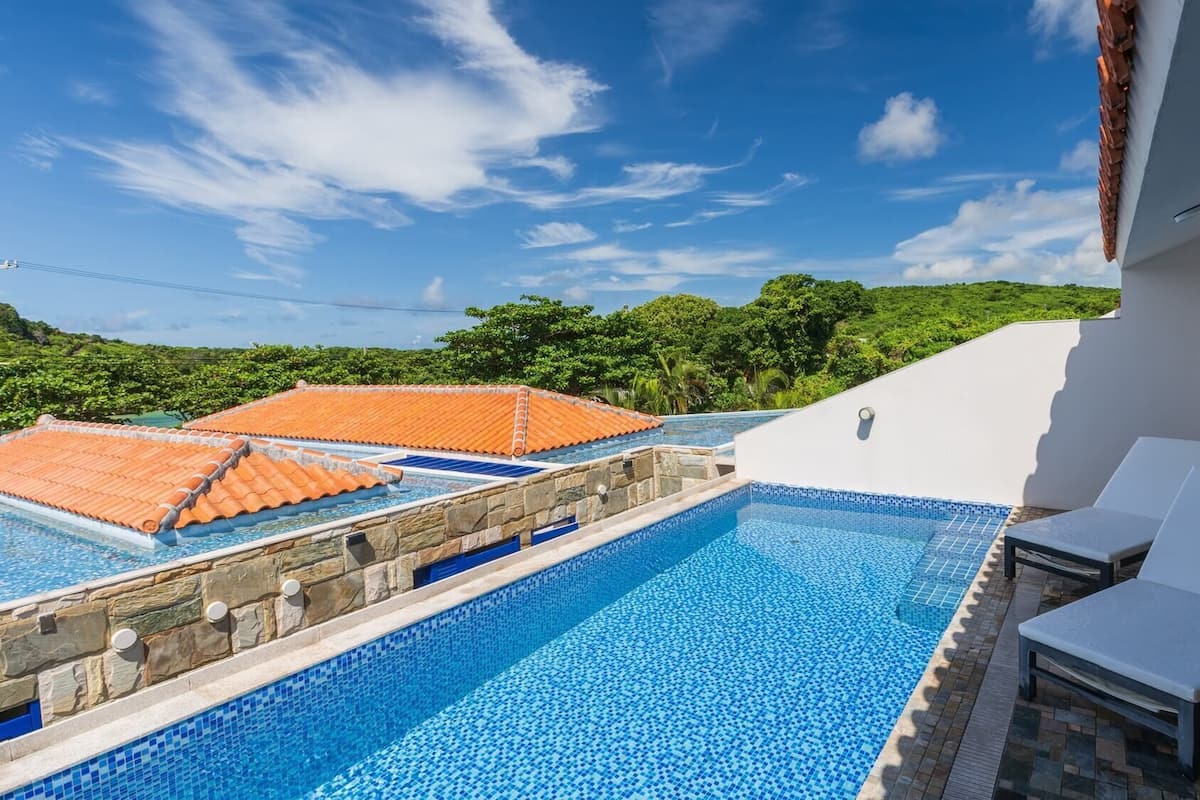 A rooftop pool with clear blue water, white lounge chairs, and a view of green hills.