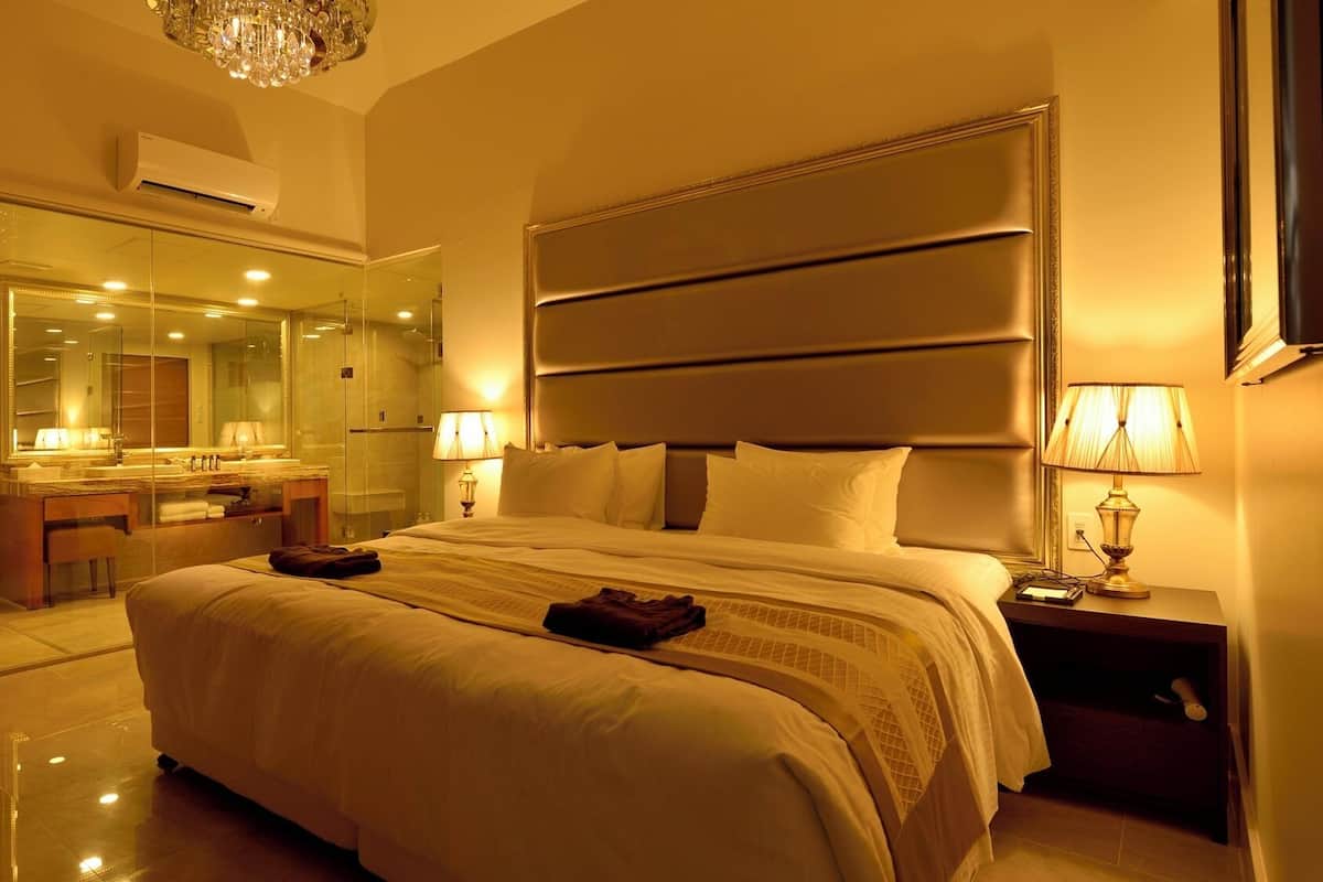 A hotel room with a large bed, a bathroom with a glass shower enclosure, a vanity with a mirror, and a chandelier.